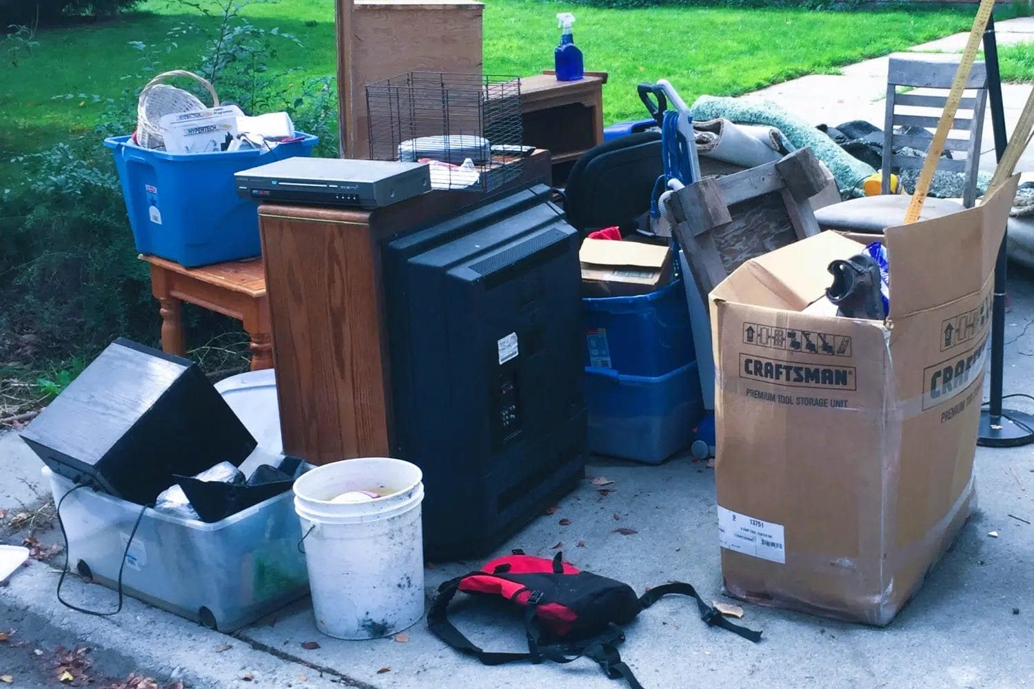 A pile of unwanted household junk including boxes, an old TV, small furniture, and storage bins left on a driveway for junk removal