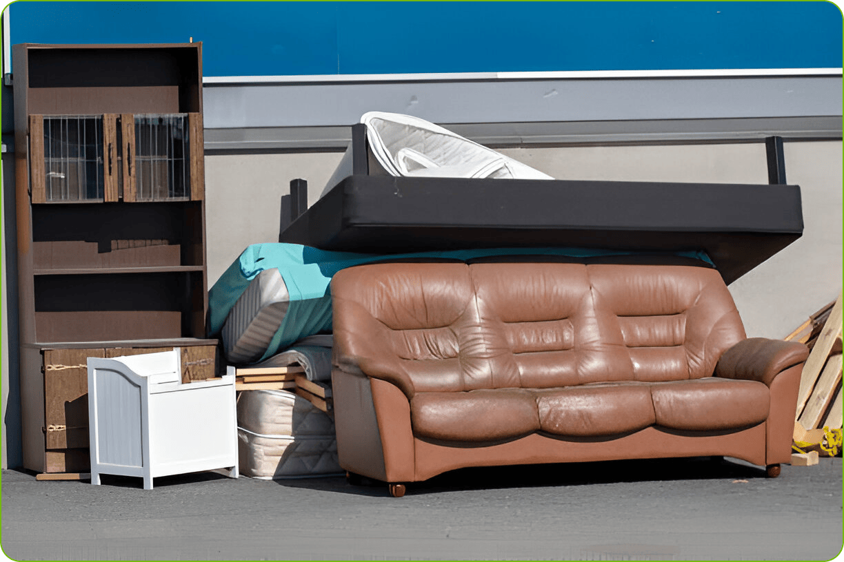 A pile of discarded household furniture including a brown leather sofa, mattresses, and shelving left outside for junk removal or disposal