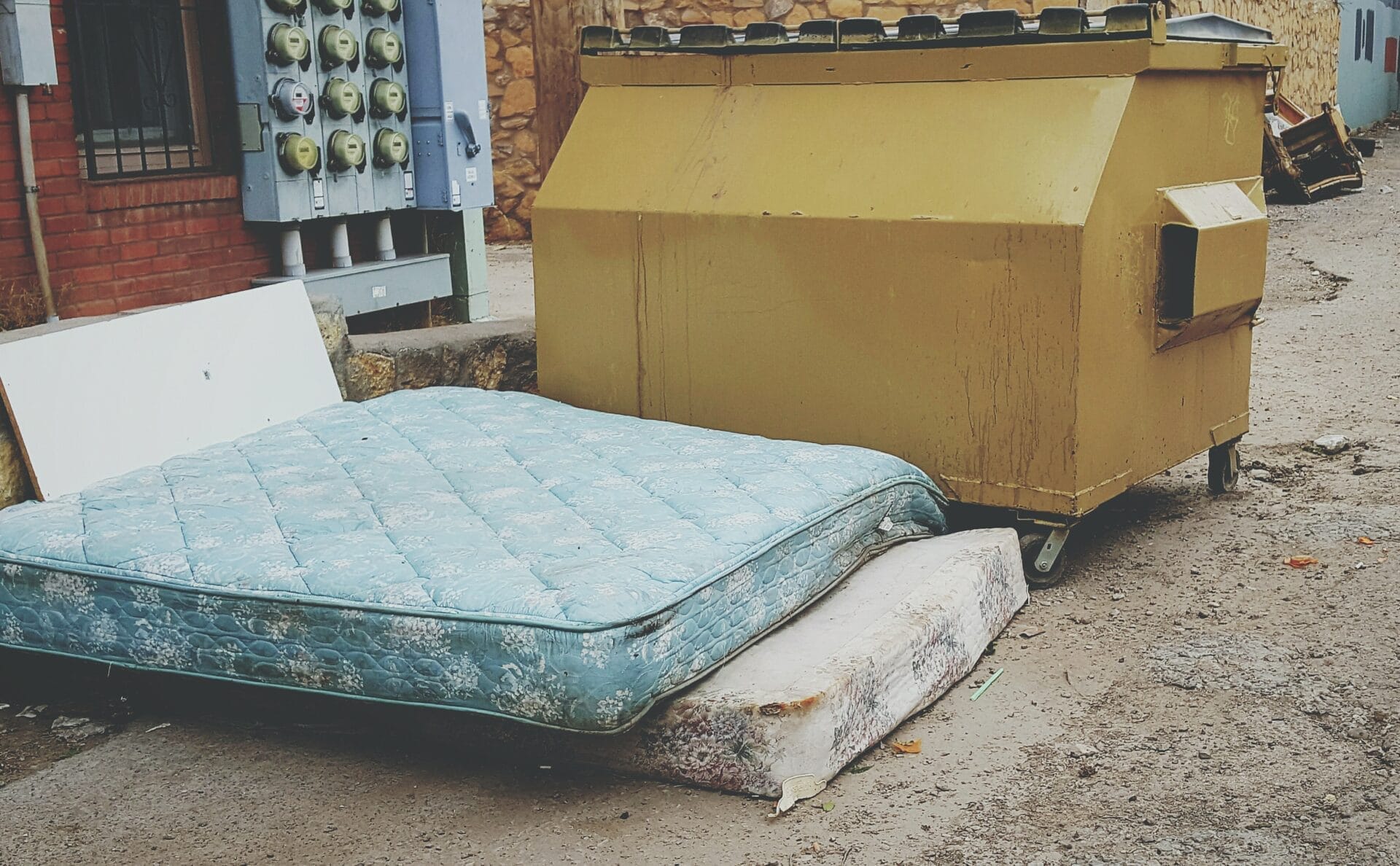 Two old mattresses discarded next to a large dumpster, awaiting pickup for junk removal
