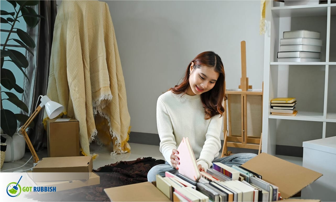 Young woman smiles while sorting books into cardboard boxes, preparing items for donation, moving, or decluttering