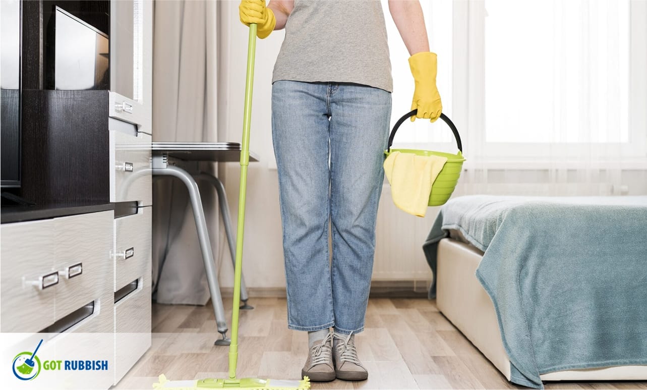 A person in yellow gloves holds a mop and a green bucket while cleaning a bright bedroom, showing a spring cleaning service