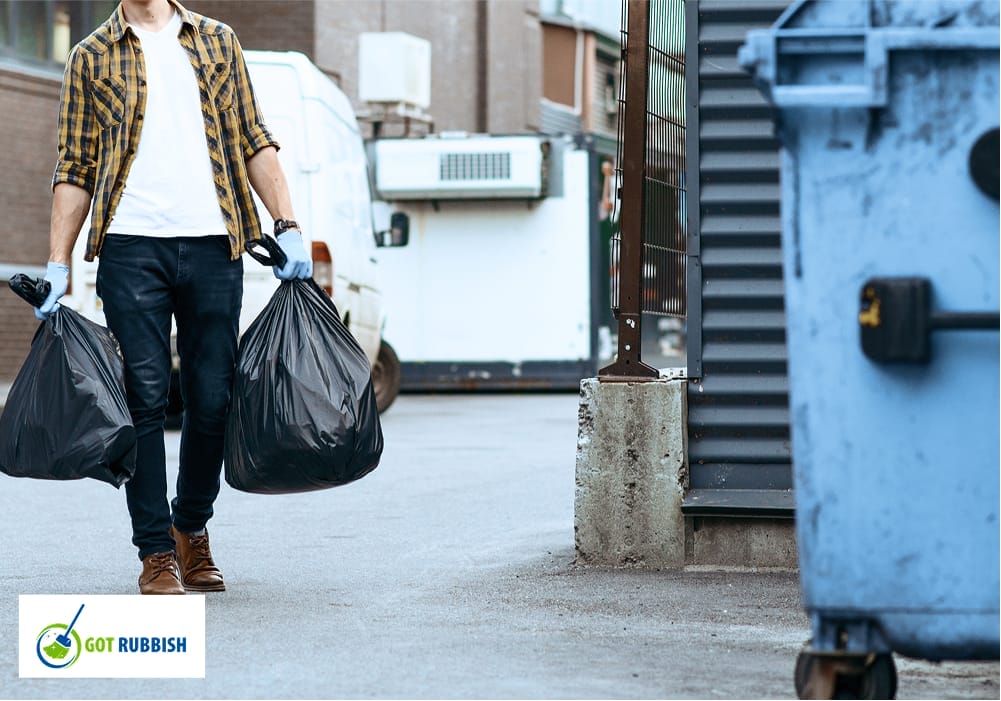 A person carrying two large black garbage bags toward a dumpster, representing waste disposal and junk hauling services in an urban alleyway