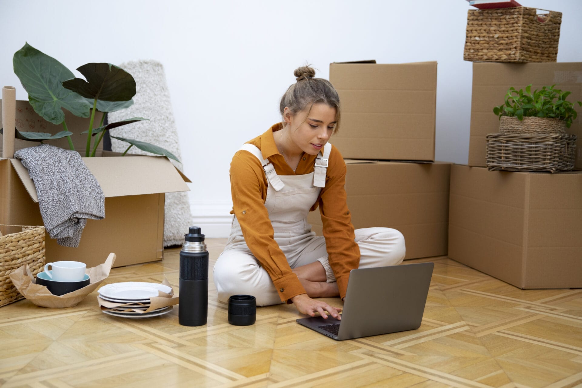 A young woman sitting cross-legged on the floor of her new home, surrounded by moving boxes, dishes, and plants, using her laptop to organize arrange home cleanout services