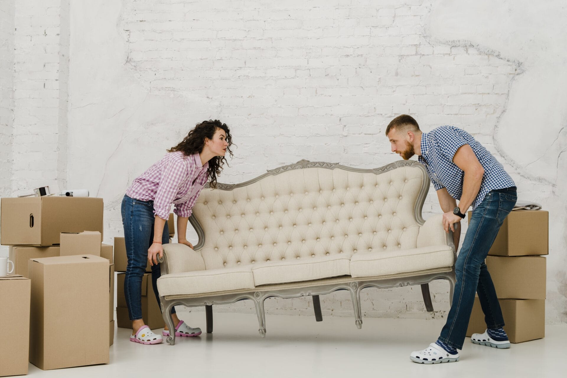 A man and woman carefully lifting a vintage cream-colored sofa with tufted upholstery while surrounded by moving boxes, symbolizing home relocation or furniture moving